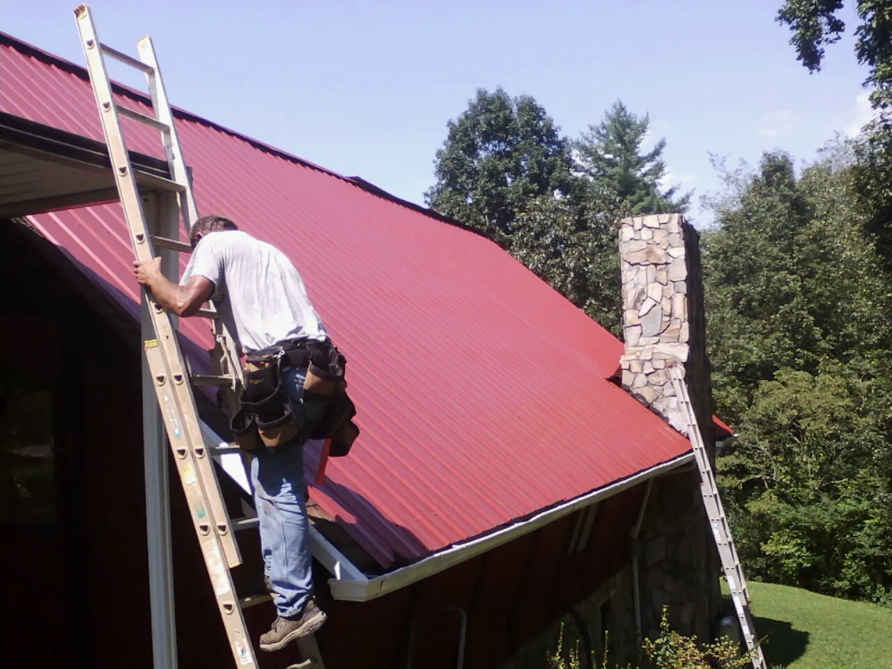 Roofer on a ladder beside a red metal roof and stone chimney — Soffit & Fascia Repair by Roofing Brothers in Weir